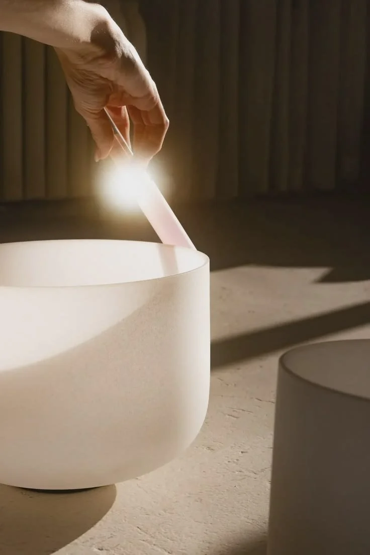 A hand lifting a wand from a quartz crystal singing bowl in soft light.